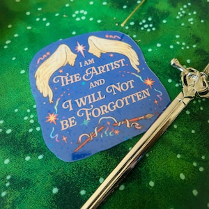 May include: A blue sticker with a pair of white wings and the text "I am the artist and I will not be forgotten". The sticker has a glittery finish and features a paintbrush and stars.