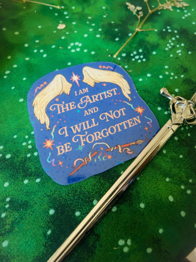 May include: A blue sticker with a pair of white wings and the text "I am the artist and I will not be forgotten". The sticker has a glittery finish and features a paintbrush and stars.