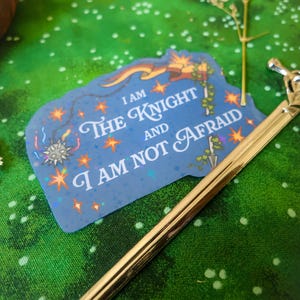 May include: A blue sticker with a glittery finish and a white border. The sticker features a knight with a sword and a banner that reads "I am the Knight and I am not afraid".