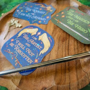 May include: Three holographic stickers with inspirational quotes. The first sticker is blue and features the text "I am the Knight and I am not afraid". The second sticker is green and features the text "I am the Maiden and I am the Hero". The third sticker is blue and features the text "I am the Artist and I will not be forgotten".
