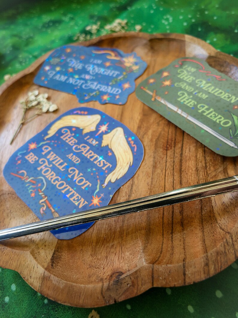 May include: Three holographic stickers with inspirational quotes. The first sticker is blue and features the text "I am the Knight and I am not afraid". The second sticker is green and features the text "I am the Maiden and I am the Hero". The third sticker is blue and features the text "I am the Artist and I will not be forgotten".