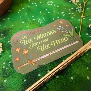 May include: A green sticker with a gold sword and the text "I am the maiden and I am the hero" with a red ribbon and white flowers.