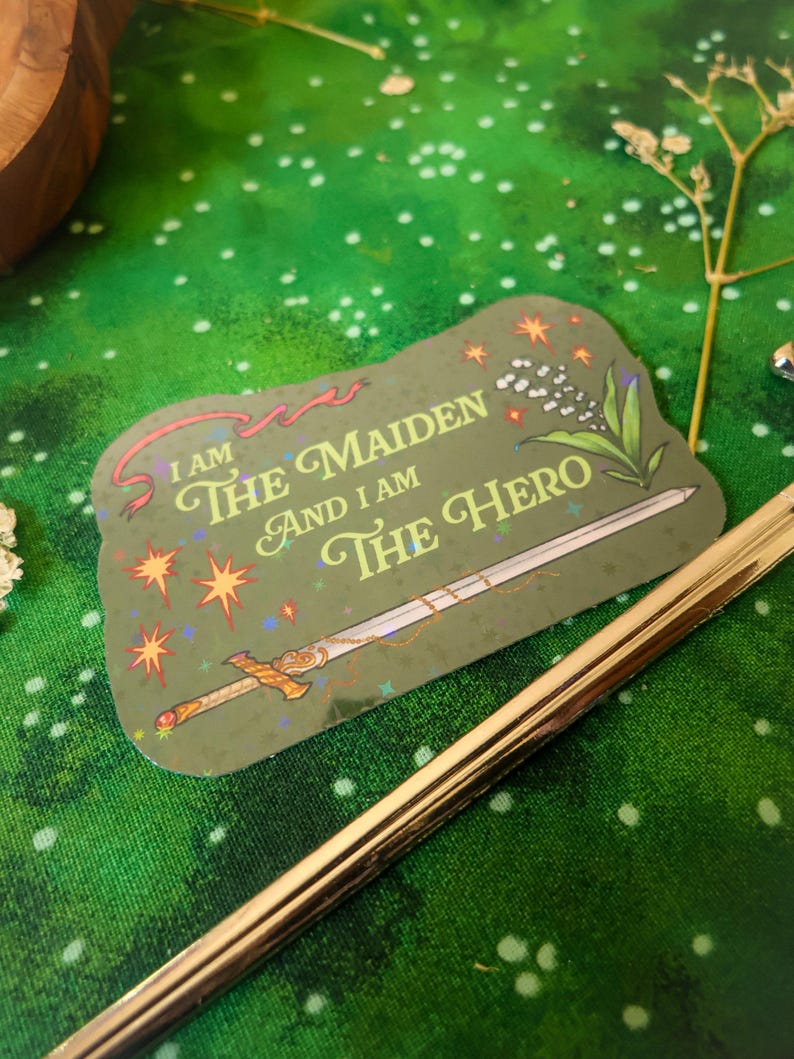 May include: A green sticker with a gold sword and the text "I am the maiden and I am the hero" with a red ribbon and white flowers.