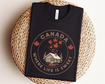 May include: Black t-shirt with a circular graphic featuring a cabin, maple leaves, and the text "CANADA WHERE LIFE IS SWEET." The design is in shades of brown and red, evoking a rustic, outdoorsy theme.