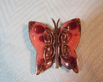 Könnte beinhalten: Eine Brosche in Form eines rosa und kupferfarbenen Schmetterlings mit filigranen Details. Der Schmetterling hat eine strukturierte Oberfläche und eine metallische Oberfläche.