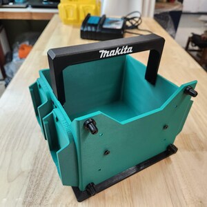 Puede incluir: Un organizador de herramientas Makita de color verde azulado y negro con un asa negra. El logotipo de Makita está impreso en blanco en el asa. El organizador tiene una base rectangular y está diseñado para guardar herramientas.