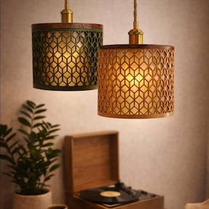 Peut inclure: Deux suspensions lumineuses avec découpes géométriques. L'une est vert olive, l'autre dorée. Les deux ont des dessus en bois et des fixations en laiton. Les lumières pendent de cordes en corde, dégageant une lueur chaleureuse.