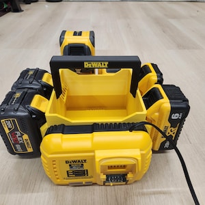 Op de afbeelding: Een geel en zwart DeWalt acculaadstation met een centrale handgreep. Het apparaat heeft meerdere sleuven met diverse 20V en 60V FlexVolt lithium-ion accu's. Een zwarte stroomkabel steekt uit de voorzijde van de acculader.