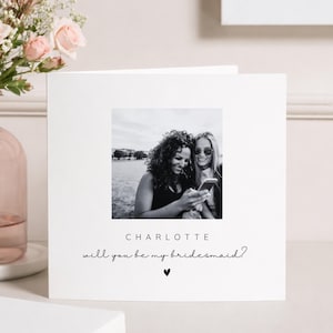 May include: A square white greeting card featuring a centred black and white photograph of two people looking at a phone. Below the photo, the text reads CHARLOTTE followed by the script text will you be my bridesmaid? and a small black heart.