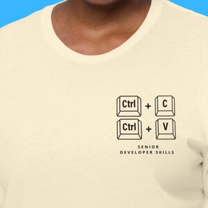 Puede incluir: Camiseta color crema con un gráfico negro de los atajos de teclado Ctrl+C y Ctrl+V. Debajo del gráfico, el texto dice "SENIOR DEVELOPER SKILLS". La camiseta tiene cuello redondo y mangas cortas.