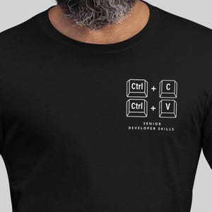 May include: Black t-shirt with a white graphic of keyboard shortcuts: Ctrl + C and Ctrl + V. The text "SENIOR DEVELOPER SKILLS" is printed below the graphic. The shirt is made of a soft material.