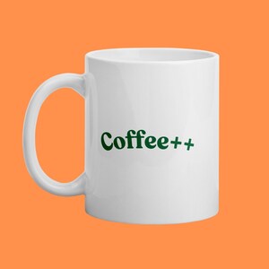 Peut inclure: Mug à café en céramique blanche avec une anse incurvée. Le mug porte l'inscription "Coffee++" en vert, dans une police stylisée. Le mug est posé sur un fond orange, créant une image à fort contraste.