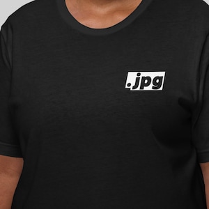 Puede incluir: Camiseta negra con un logotipo rectangular blanco que dice ".jpg". La camiseta tiene cuello redondo y mangas cortas. El logotipo está situado en la parte superior izquierda del pecho.