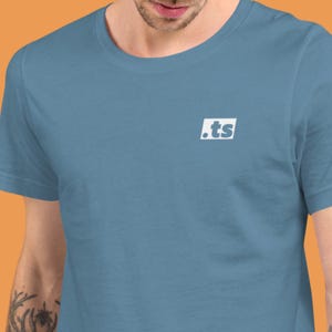 Könnte beinhalten: Hellblaues T-Shirt mit einem weißen quadratischen Logo mit der Aufschrift ".ts". Das Shirt besteht aus einem weichen Material und hat einen Rundhalsausschnitt. Das Bild zeigt den Oberkörper einer Person, die das T-Shirt trägt.