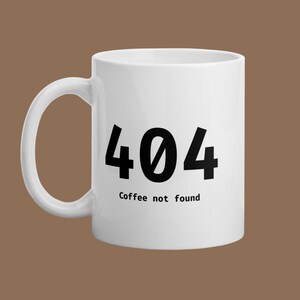 Puede incluir: Taza de café de cerámica blanca con asa negra. La taza presenta el número "404" en negrita negra, con el texto "Coffee not found" debajo. La taza está sobre un fondo marrón.