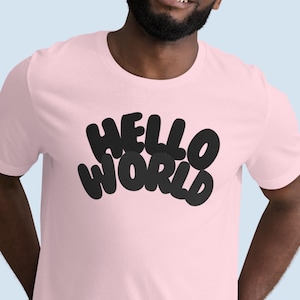 May include: A light pink t-shirt with the words "HELLO WORLD" in large, black, bubble letters. The shirt is short-sleeved and has a crew neck. The text is centered on the front of the shirt.