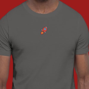 May include: A dark grey t-shirt with a small, pixelated rocket design in red, orange, and blue on the upper chest. The rocket has a flame coming out of the bottom. The shirt is made of a soft material.
