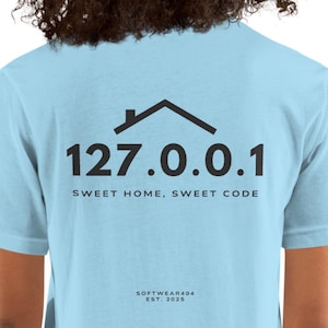 Puede incluir: Camiseta azul claro con un gráfico negro de una casa sobre los números "127.0.0.1". Debajo de los números está el texto "SWEET HOME, SWEET CODE". El texto "SOFTWARE404 EST. 2025" está en la parte inferior.