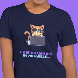 以下が含まれることがあります： メガネをかけたオレンジ色の猫がノートパソコンを使っている漫画のネイビーブルーのTシャツ。「PURRGRAMMING IN PROGRESS...」の文字が紫とターコイズで下に印刷されています。