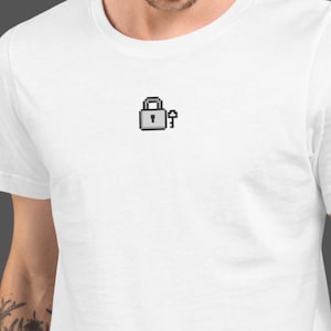 Puede incluir: Camiseta blanca con un diseño pixelado de candado y llave en tonos grises. El gráfico está situado en la parte superior del pecho. La camiseta está hecha de un material suave y cómodo.