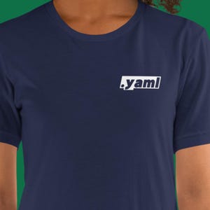 Puede incluir: Camiseta azul marino con la palabra ".yaml" en blanco, dentro de un rectángulo blanco. La camiseta es de color liso y tiene cuello redondo. La imagen muestra la parte superior del cuerpo de una persona que lleva la camiseta.