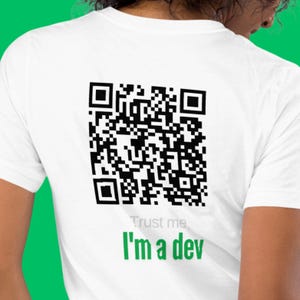 Puede incluir: Camiseta blanca con un gran código QR negro impreso en la espalda. Debajo del código, el texto "Trust me, I'm a dev" está escrito en verde. Una camiseta sencilla e informal.