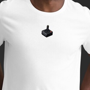 May include: White t-shirt featuring a pixelated black joystick design on the chest. The joystick has a red button and a black handle with a gold tip. The shirt is made of a soft, comfortable material.