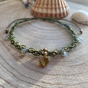 May include: A handmade macrame bracelet in olive green with small gold beads and light blue stones. A gold leaf charm hangs from the center. The bracelet is displayed on a wooden surface with seashells in the background.