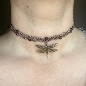 May include: A taupe macrame choker necklace featuring a central bronze-toned dragonfly pendant. The band is woven with small metallic beads and interspersed with round, brown, semi-precious stone beads along the entire length of the cord.
