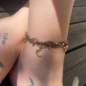 Op de afbeelding: Bruin macramé enkelbandje met goudkleurige accenten, waaronder kleine kralen en een maanvormige bedel. De armband wordt om een enkel gedragen, waarbij het ontwerp en de textuur van de sieraden worden getoond.