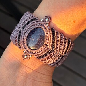 May include: A wide macrame bracelet featuring a central oval blue goldstone cabochon with shimmering flecks. The bracelet is crafted from mauve and black cord, incorporating intricate knot patterns and small metallic silver beads as decorative accents.