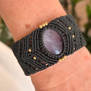 May include: A wide black macrame bracelet featuring a central oval-shaped purple amethyst gemstone. The dark cord is intricately knotted in a geometric pattern and accented with small, round, metallic gold-coloured beads embedded throughout.