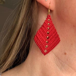 May include: Red macrame earrings with gold beads. The diamond-shaped earrings have a woven design. They hang from gold-coloured ear wires. These earrings are a striking accessory.