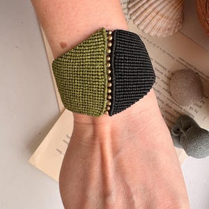 May include: A wide macrame bracelet in olive green and black, with a row of small gold beads down the centre. The bracelet is on a wrist, with a background of a book and seashells.