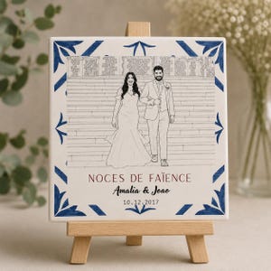 May include: A square ceramic tile featuring a line drawing of a couple on stairs, framed by a blue and white geometric border. The text reads NOCES DE FAÏENCE, Amalia & Joao, and 10.12.2017. The tile is displayed on a small wooden easel stand.