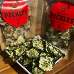May include: A white square dish filled with dried cucumber slices, seasoned with herbs. Two clear bags of Bunny Pickles are in the background, each with a red label. The label reads "Bunny Pickles" in white text.