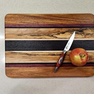 May include: A rectangular wooden cutting board with a striped design. The board has alternating bands of light and dark wood, with purple and black accents. A small knife and a red apple are on the board.