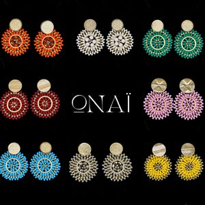 Pode incluir: Uma coleção de brincos de contas em várias cores, incluindo laranja, branco, verde, vermelho, rosa, azul, cinza e amarelo. Cada par apresenta um disco dourado no topo e um design circular de contas. A palavra "ONAÏ" é exibida em branco.