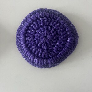 Puede incluir: Un estropajo circular de color morado intenso hecho de hilo grueso y texturizado. El artículo presenta un patrón de ganchillo denso en espiral que irradia desde el centro hacia los bordes, creando una forma redondeada firme con un acabado brillante.