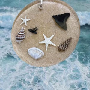 May include: A round, sand-colored resin ornament with a silver string for hanging. The ornament is decorated with seashells, white starfish, and black shark teeth. The background features a blurred image of ocean waves.