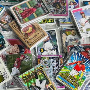 May include: A collection of baseball trading cards, many in protective sleeves. The cards feature various players and teams, including the Yankees, Dodgers, and Angels. Some cards have the text "2021's Latest Hits".