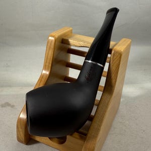 Könnte beinhalten: Eine mattschwarze Tabakpfeife mit gebogenem Mundstück, die auf einem hellen Pfeifenständer aus Holz ruht. Die Pfeife hat ein silbernes Band in der Nähe der Verbindung des Mundstücks mit dem Kopf. Der Ständer ist aus hellem Holz.