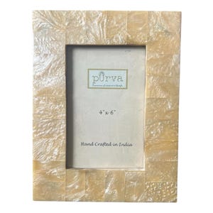 May include: Rectangular photo frame with a mosaic-like design. The frame is made of small, rectangular pieces in shades of cream and beige. The frame is labeled "4" x 6"" and "Hand Crafted in India". The brand name "pürva" is also visible.