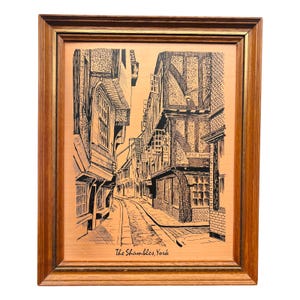 May include: Framed copper etching of The Shambles in York, England. The artwork depicts a narrow cobblestone street lined with historic buildings. The frame is a warm brown with a gold inner edge, enhancing the vintage aesthetic.
