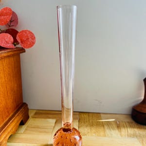 May include: A clear glass bud vase with a round, textured base and a long, slender neck. The base is a warm amber color, and the vase stands on a wooden surface. The vase is designed to hold a single flower or small floral arrangement.