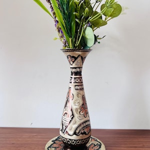 May include: A tall, slender metal vase with a flared rim, featuring intricate black, copper, and cream-colored etched patterns. The vase sits on a matching circular plate, and it is filled with a bouquet of green, purple, and white artificial flowers.