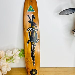 May include: A wooden decorative item with a long, oval shape. It features a painted design in black, yellow, and white, with a stylized image of a weapon. The item has a sticker that says "Australian Made". The bottom has a sticker that says "100% Aboriginal Owned".