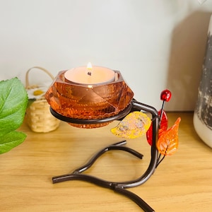 Op de afbeelding: Een decoratieve kandelaar met een brandende theelichtkaars. De houder heeft een oranje, facetgeslepen glazen beker en een donkerbruine metalen basis met bladeren. De kandelaar is ontworpen om een enkele theelichtkaars te bevatten.
