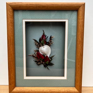 Può includere: Opera d'arte incorniciata con una conchiglia a forma di cuore e boccioli di rose rosse. L'opera è ambientata su uno sfondo azzurro chiaro all'interno di un passe-partout bianco, racchiusa in una cornice di legno. La cornice ha una finitura a venatura naturale.