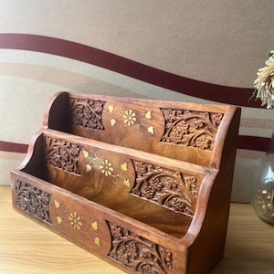 May include: A wooden desk organizer with three tiered compartments. The organizer is a warm brown color with intricate floral carvings and gold accents. The design is symmetrical, with detailed patterns on the front and sides.